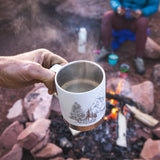 Close up of a hand holding the Klean Kanteen eco-friendly metal travel mug over a camp fire