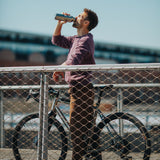 A man standing over a bike on a bridge and drinking from a Klean Kanteen stainless steel insulated reflect rise insulated bottle