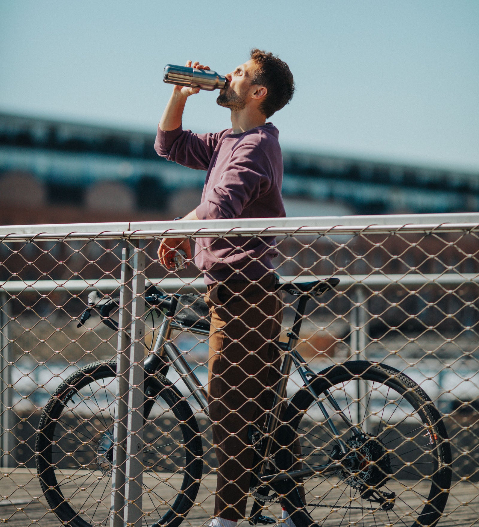A man standing over a bike on a bridge and drinking from a Klean Kanteen stainless steel insulated reflect rise insulated bottle