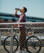 A man standing over a bike on a bridge and drinking from a Klean Kanteen stainless steel insulated reflect rise insulated bottle