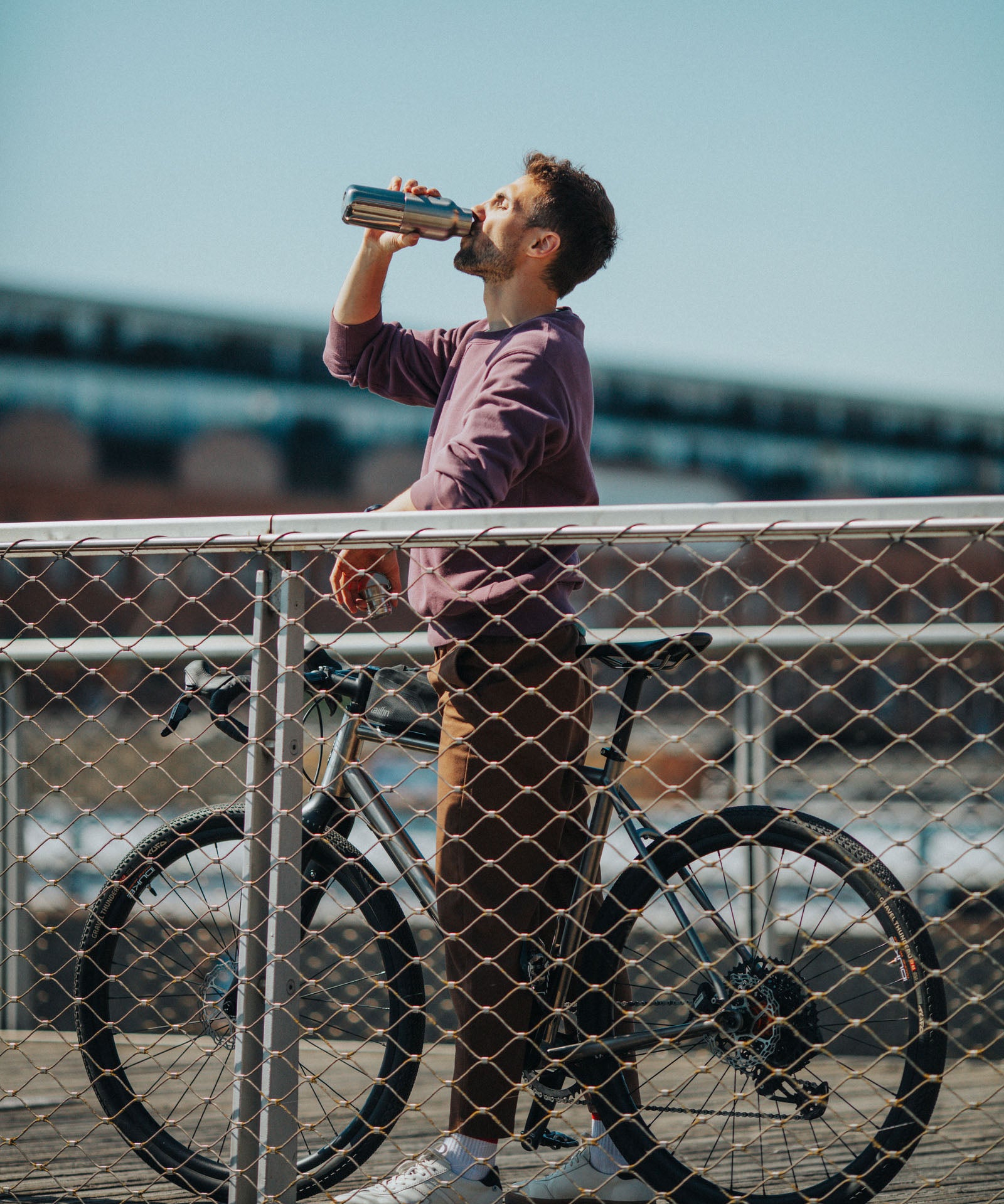A man standing over a bike on a bridge and drinking from a Klean Kanteen stainless steel insulated reflect rise insulated bottle