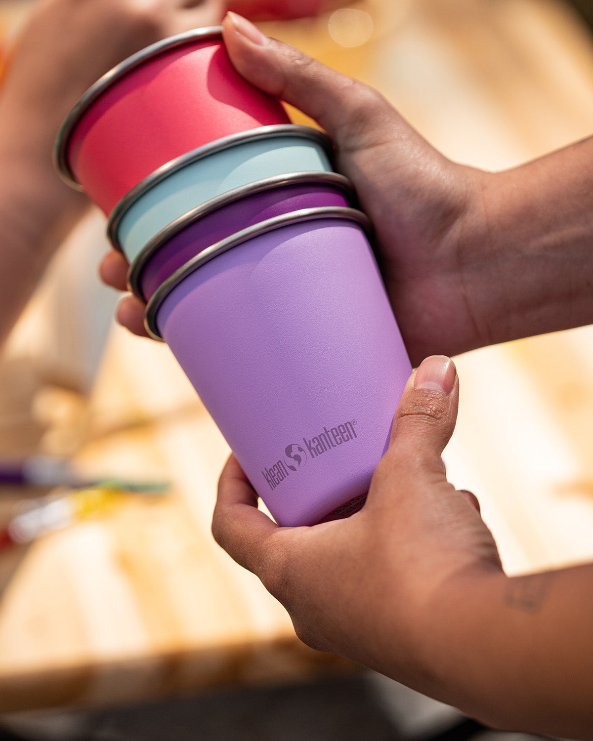 Hands holding a stack of Klean Kanteen stainless steel kid cups 10oz in bright pinks and purples - plastic free, made from post-consumer steel, available to buy at Babipur.