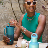 A woman sitting at a table smiling with a brittany blue coloured 26oz handled Klean Kanteen insulated tumbler in her hands.