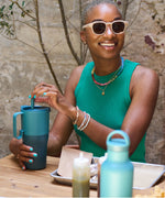 A woman sitting at a table smiling with a brittany blue coloured 26oz handled Klean Kanteen insulated tumbler in her hands.