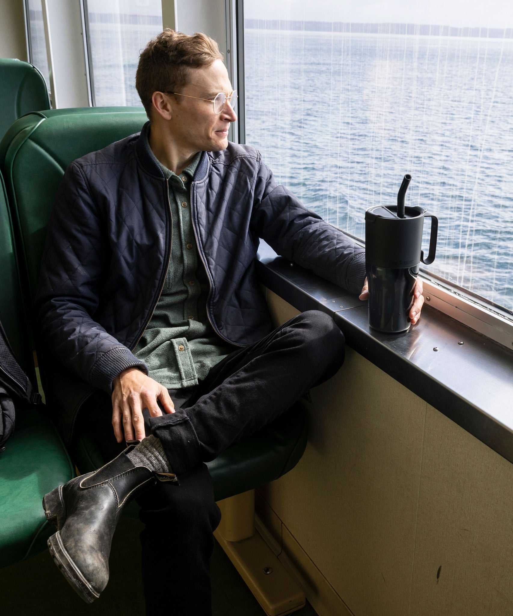 A man sitting by a window on a ferry. He is holding the base of a black coloured 26oz Klean Kanteen rise handled tumbler which is resting on the windowsill.