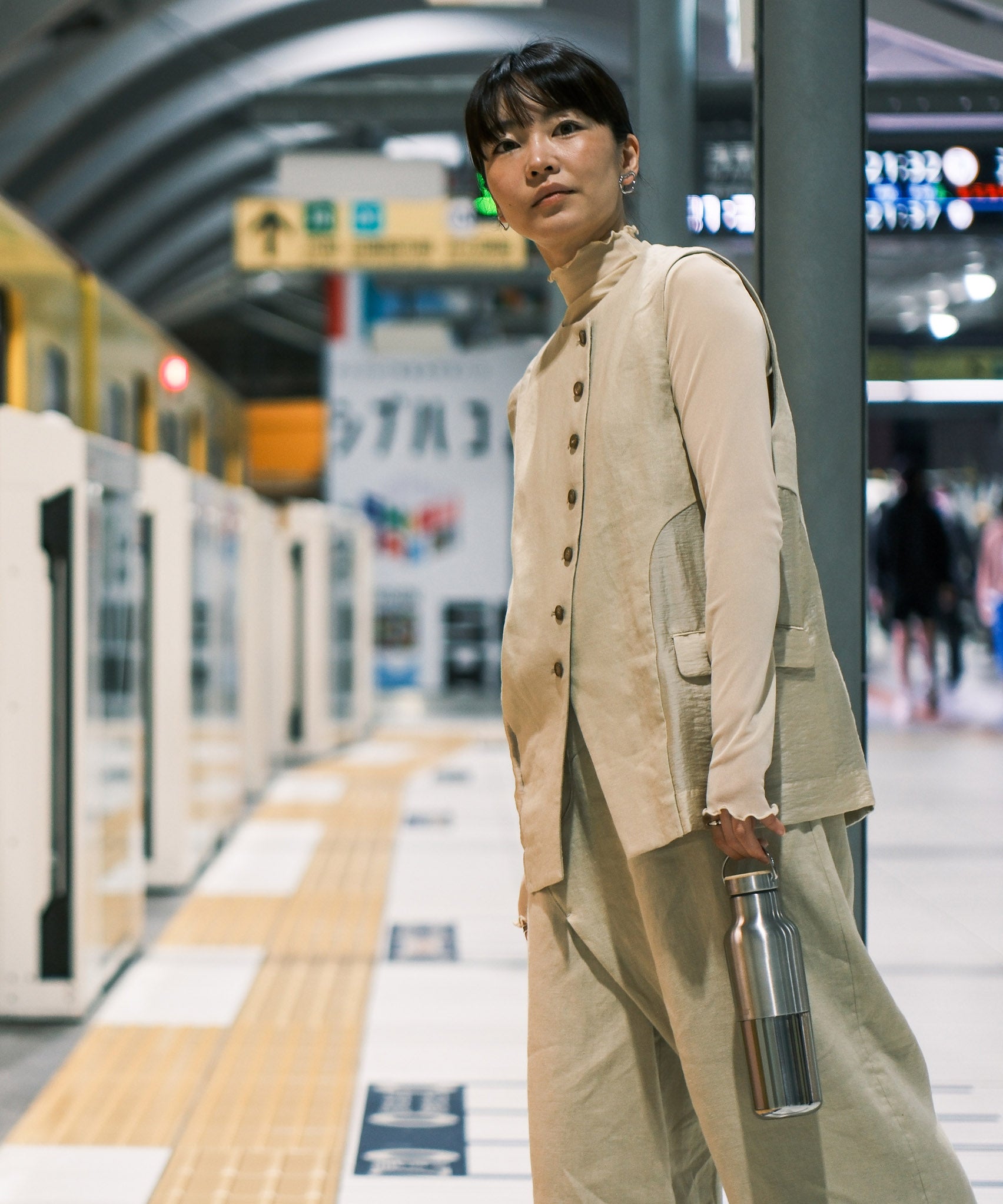 A woman standing on a platform in a train station and holding the  klean Kanteen reflect stainless steel rise bottle by the handle on the bamboo loop cap