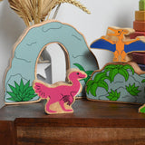 A Lanka Kade caudipteryx pink dinosaur wooden toy figure placed on a small wooden table with scenery pieces from the prehistoric play set in the background. These wooden toy figures are part of a huge range of Lanka Kade fair trade, wooden toys available here at Babipur.