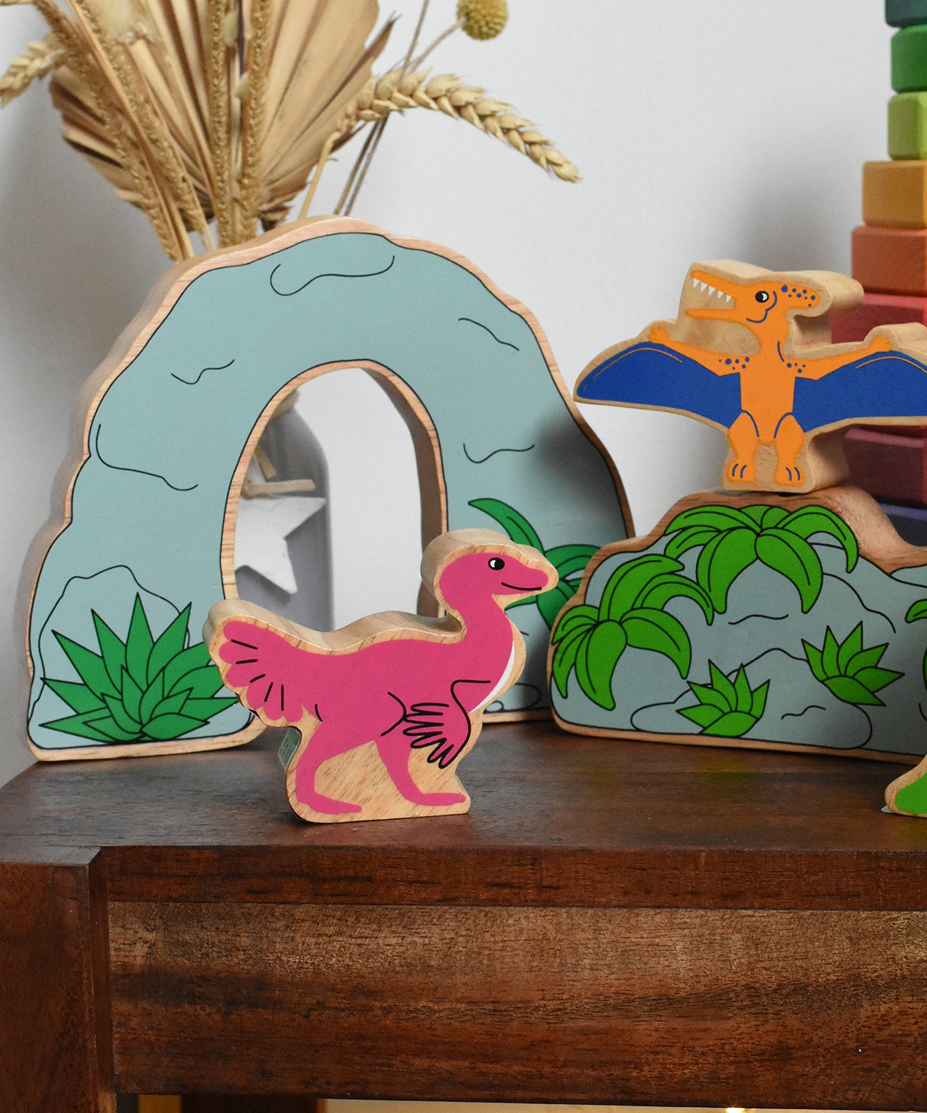 A Lanka Kade caudipteryx pink dinosaur wooden toy figure placed on a small wooden table with scenery pieces from the prehistoric play set in the background. These wooden toy figures are part of a huge range of Lanka Kade fair trade, wooden toys available here at Babipur.