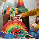 A child reaching for a Lanka Kade caudipteryx pink dinosaur wooden toy figure. These wooden toy figures are part of a huge range of Lanka Kade fair trade, wooden toys available here at Babipur.