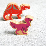 A Lanka Kade caudipteryx pink dinosaur wooden toy figure placed on carpet in front of a red spinosaurus. These wooden toy figures are part of a huge range of Lanka Kade fair trade, wooden toys available here at Babipur.