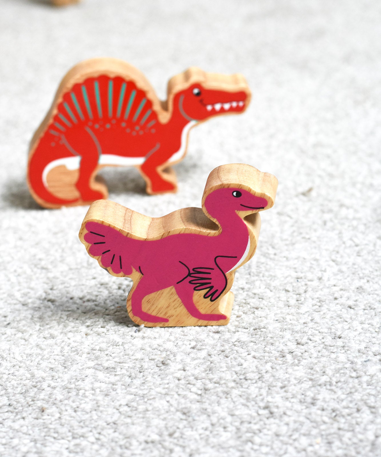 A Lanka Kade caudipteryx pink dinosaur wooden toy figure placed on carpet in front of a red spinosaurus. These wooden toy figures are part of a huge range of Lanka Kade fair trade, wooden toys available here at Babipur.