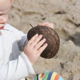 A young child pouring sand from a Lanka Kade coconut bowl on a beach. The Lanka Kade coconut collection is part of our wide range of outdoor toys here at Babipur.
