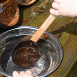 A close up of a child using the Lanka Kade coconut shell spoon utensil. The child is scooping water out of a small water bowl.