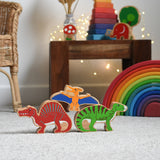 Lanka Kade wooden toy dinosaur figures placed on carpet. A yellow pteranodon figure has been balanced on a red spinosaurus and a green iguanodon. These wooden toy figures are part of a huge range of Lanka Kade fair trade, wooden toys available here at Babipur.
