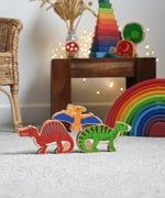 Lanka Kade wooden toy dinosaur figures placed on carpet. A yellow pteranodon figure has been balanced on a red spinosaurus and a green iguanodon. These wooden toy figures are part of a huge range of Lanka Kade fair trade, wooden toys available here at Babipur.