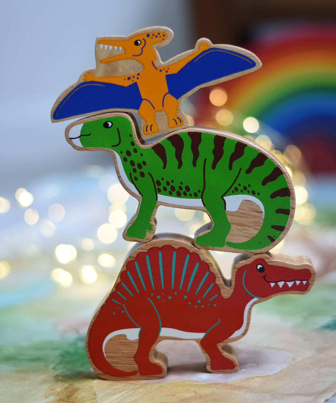 A stack of three wooden dinosaur figures from Lanka Kade. A yellow pteranodon. green iguanodon and red spinosaurus. These wooden toy figures are part of a huge range of Lanka Kade fair trade, wooden toys available here at Babipur.