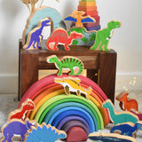 A large collection of Lanka Kade wooden toy dinosaur figures placed on a small wooden table and a Grimm's rainbow. These wooden toy figures are part of a huge range of Lanka Kade fair trade, wooden toys available here at Babipur.