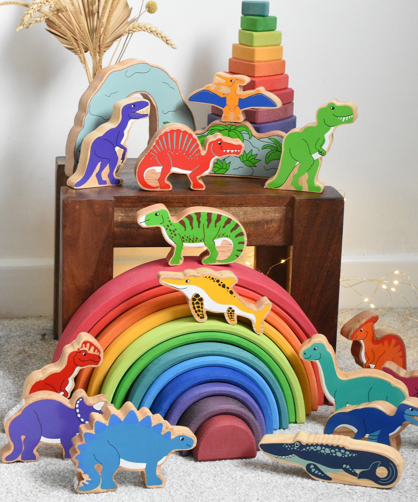 A large collection of Lanka Kade wooden toy dinosaur figures placed on a small wooden table and a Grimm's rainbow. These wooden toy figures are part of a huge range of Lanka Kade fair trade, wooden toys available here at Babipur.