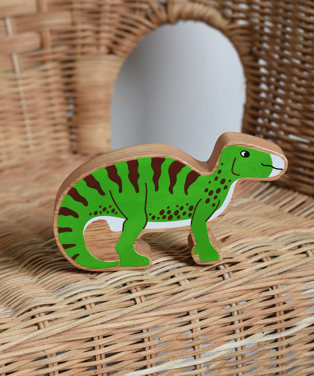 A Lanka Kade green iguanodon wooden toy figure placed on a child's wicker chair. These wooden toy figures are part of a huge range of Lanka Kade fair trade, wooden toys available here at Babipur.
