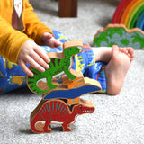 A stack of three wooden dinosaur figures from Lanka Kade. A yellow pteranodon. green iguanodon and red spinosaurus. A child's hand can be seen holding the iguanadon toy.These wooden toy figures are part of a huge range of Lanka Kade fair trade, wooden toys available here at Babipur.