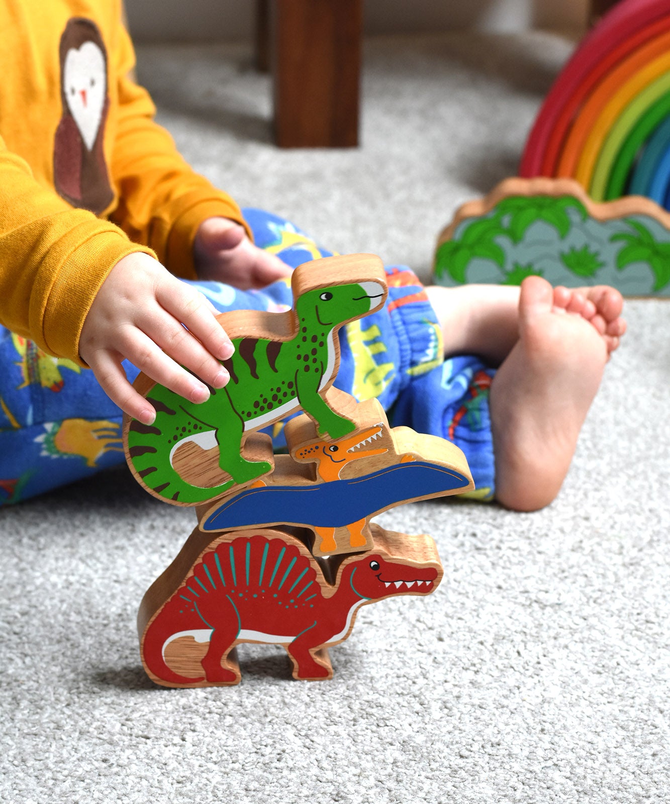 A stack of three wooden dinosaur figures from Lanka Kade. A yellow pteranodon. green iguanodon and red spinosaurus. A child's hand can be seen holding the iguanadon toy.These wooden toy figures are part of a huge range of Lanka Kade fair trade, wooden toys available here at Babipur.