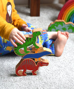 A stack of three wooden dinosaur figures from Lanka Kade. A yellow pteranodon. green iguanodon and red spinosaurus. A child's hand can be seen holding the iguanadon toy.These wooden toy figures are part of a huge range of Lanka Kade fair trade, wooden toys available here at Babipur.