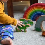 A child reaching for the Lanka Kade green iguanodon wooden toy figure, the cave and scenery pieces from the prehstoric play set can be seen n the background. These wooden toy figures are part of a huge range of Lanka Kade fair trade, wooden toys available here at Babipur.