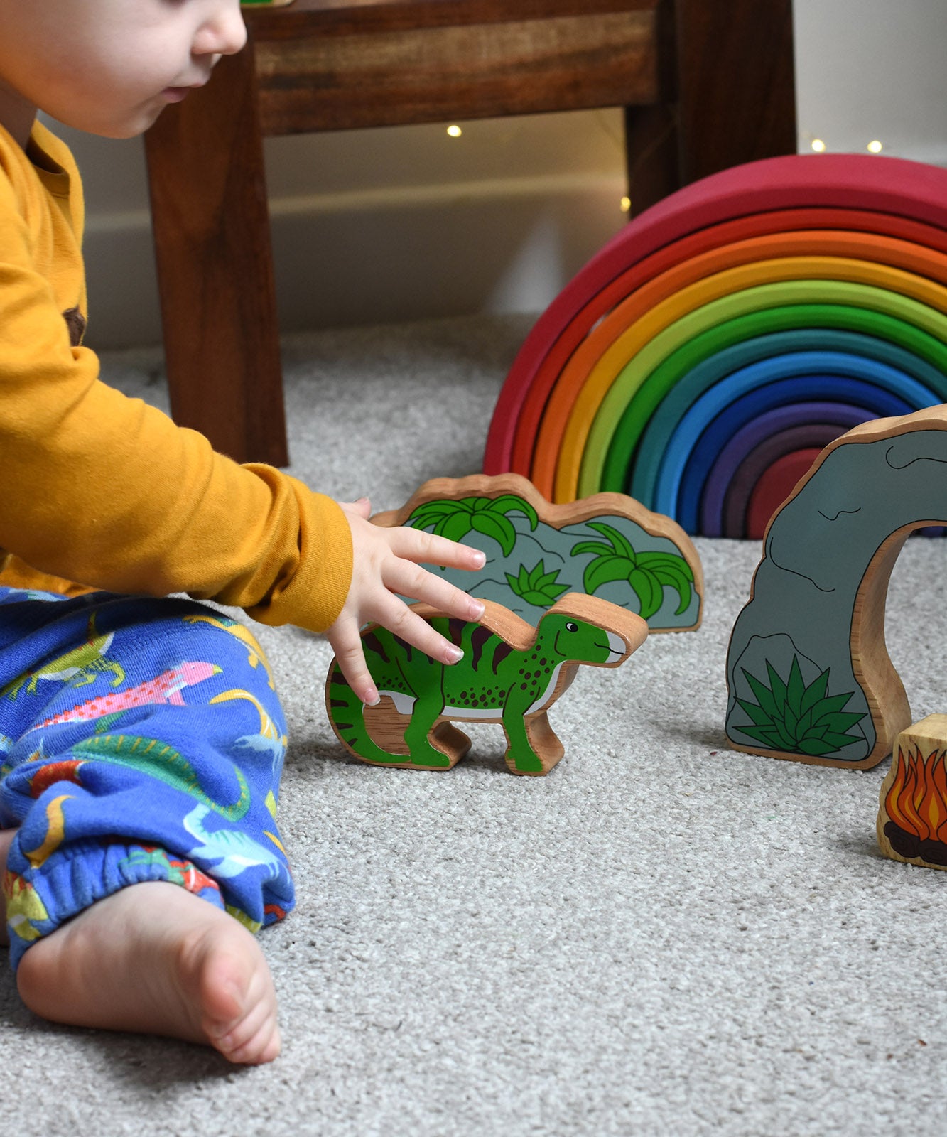 A child reaching for the Lanka Kade green iguanodon wooden toy figure, the cave and scenery pieces from the prehstoric play set can be seen n the background. These wooden toy figures are part of a huge range of Lanka Kade fair trade, wooden toys available here at Babipur.
