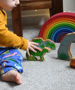 A child reaching for the Lanka Kade green iguanodon wooden toy figure, the cave and scenery pieces from the prehstoric play set can be seen n the background. These wooden toy figures are part of a huge range of Lanka Kade fair trade, wooden toys available here at Babipur.