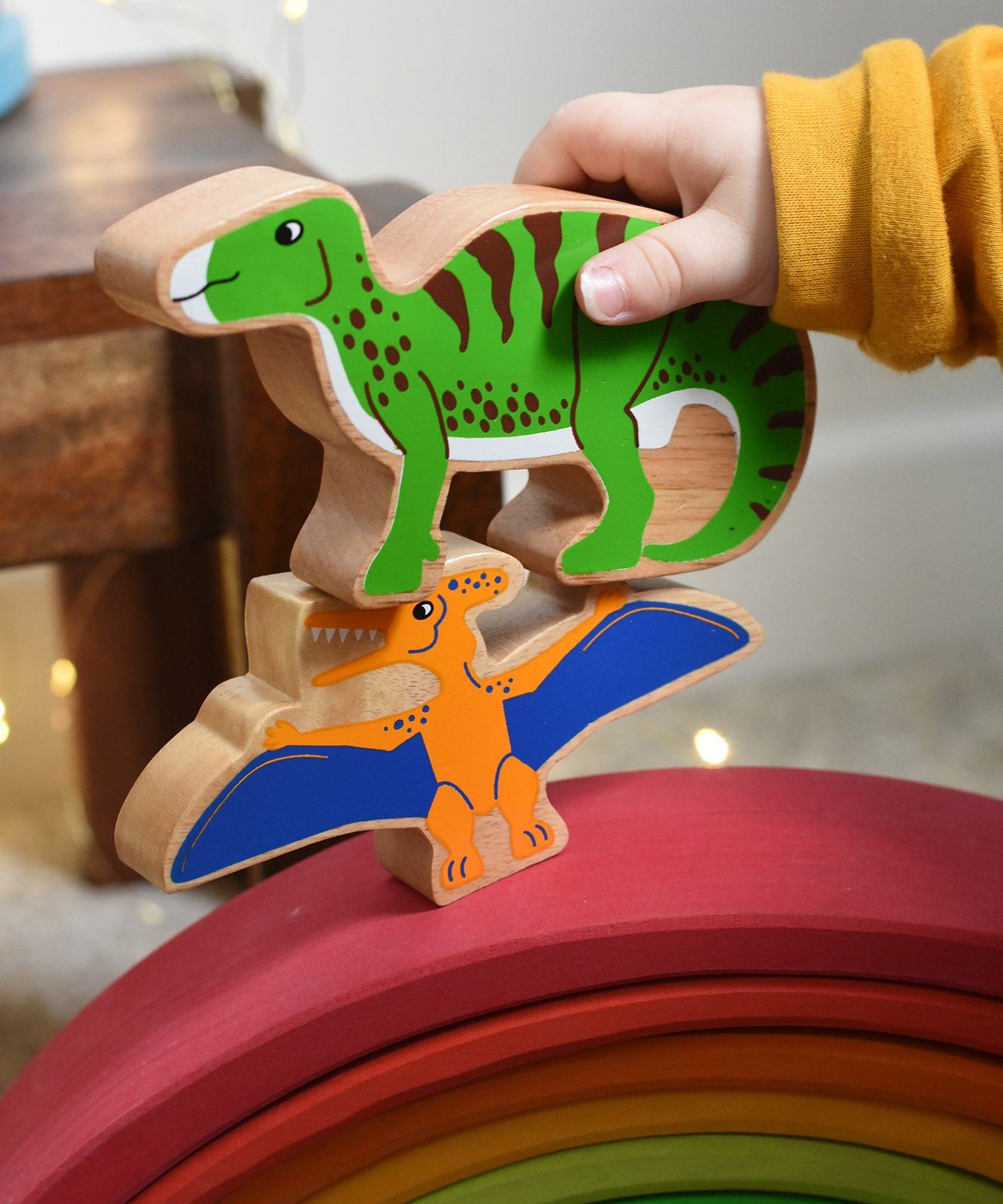 A close up of a child's hand holding the Lanka Kade green iguanodon wooden toy figure, placing it on a pteranodon figure. These wooden toy figures are part of a huge range of Lanka Kade fair trade, wooden toys available here at Babipur.