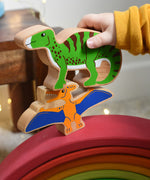 A close up of a child's hand holding the Lanka Kade green iguanodon wooden toy figure, placing it on a pteranodon figure. These wooden toy figures are part of a huge range of Lanka Kade fair trade, wooden toys available here at Babipur.