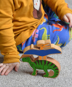 A close up of child playing with Lanka Kade wooden toy dinosaur figures. The child is pointing at the Lanka Kade pteranodon figure that has been stacked n top of a iguanodon dinosaur figure. These wooden toy figures are part of a huge range of Lanka Kade fair trade, wooden toys available here at Babipur.