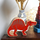 A Red Lanka Kade Wooden Spinosaurus Dinosaur placed on a small wooden table. These wooden toy figures are part of a huge range of Lanka Kade fair trade, wooden toys available here at Babipur.
