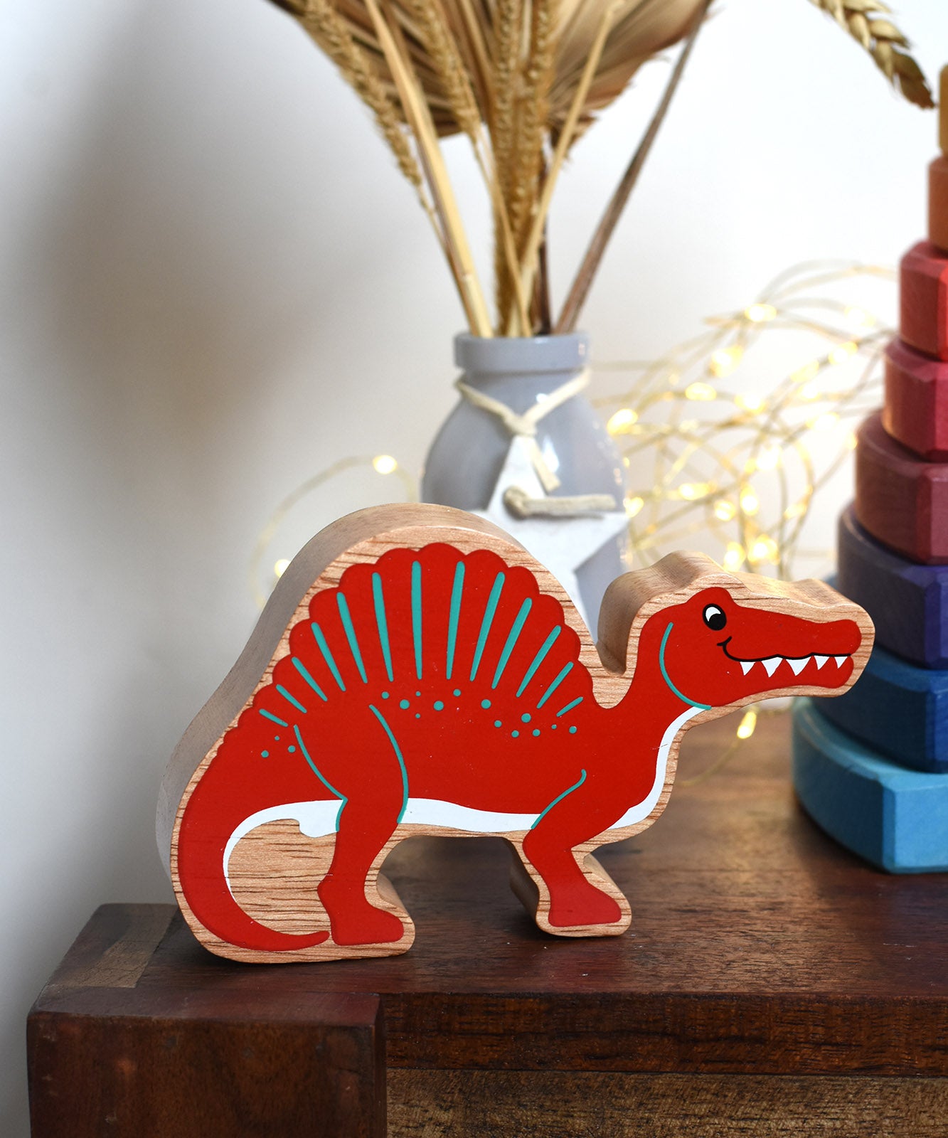 A Red Lanka Kade Wooden Spinosaurus Dinosaur placed on a small wooden table. These wooden toy figures are part of a huge range of Lanka Kade fair trade, wooden toys available here at Babipur.
