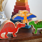 A collection of three Lanka Kade Wooden dinosaur figures placed on a small wooden table. A red Spinosaurus, a yellow pteranodon and a green iguanodon. These wooden toy figures are part of a huge range of Lanka Kade fair trade, wooden toys available here at Babipur.
