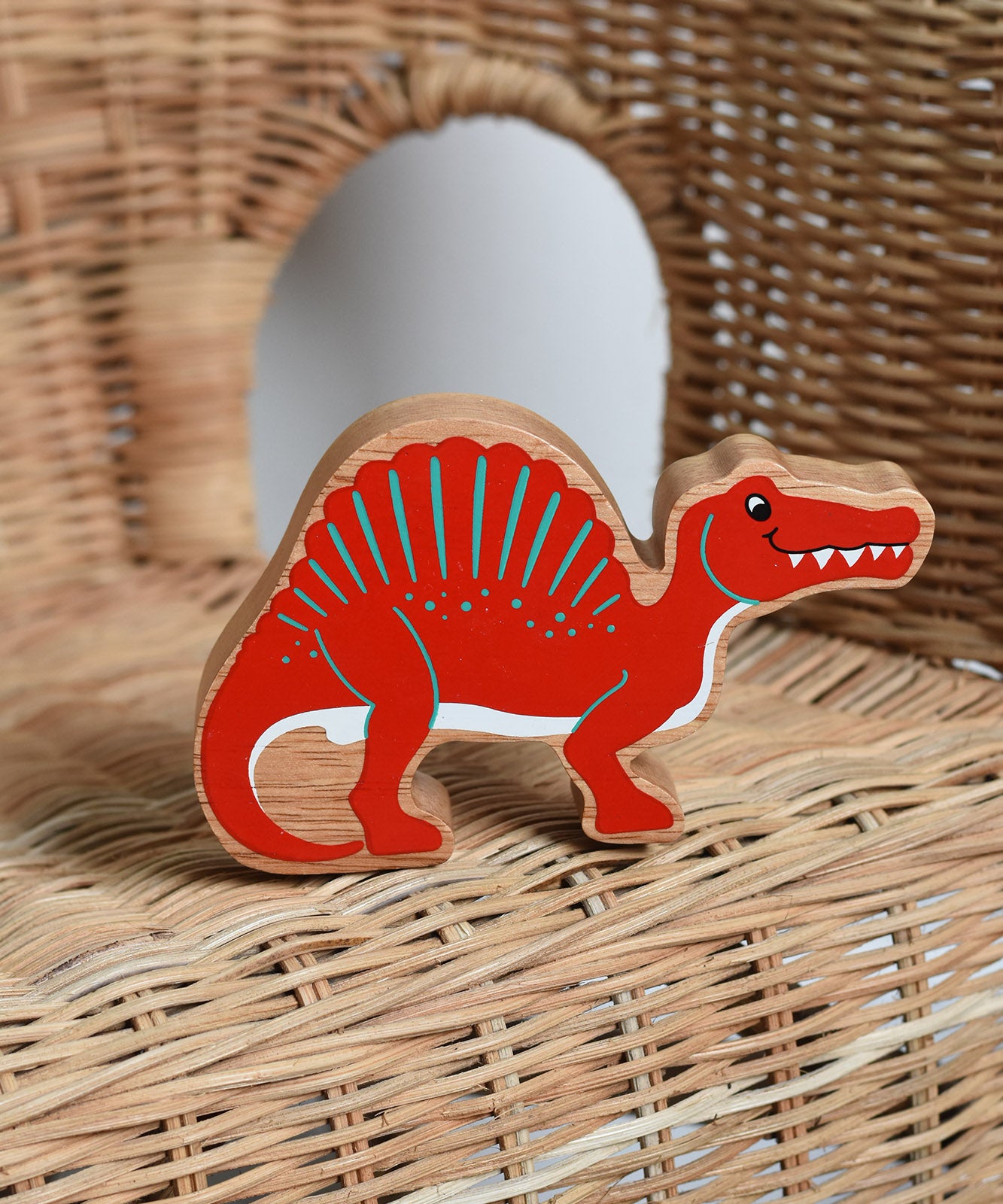 A red Lanka Kade Wooden Spinosaurus Dinosaur placed on a child's wicker chair. These wooden toy figures are part of a huge range of Lanka Kade fair trade, wooden toys available here at Babipur.