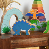 Lanka Kade Stegosaurus Dinosaur toy figure placed on a small wooden table with scenery pieces from the prehistoric play set in the background. These wooden toy figures are part of a huge range of Lanka Kade fair trade, wooden toys available here at Babipur.