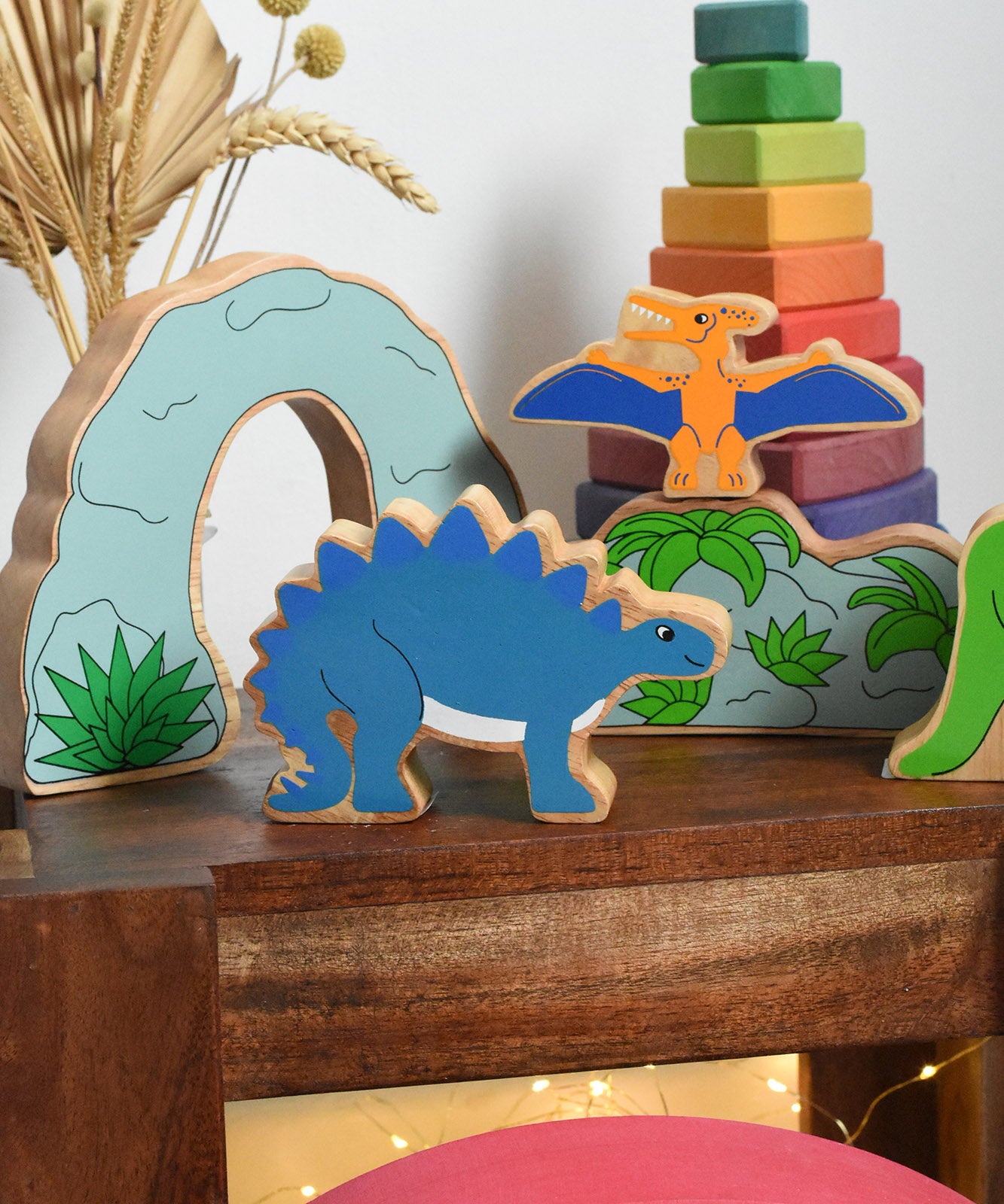 Lanka Kade Stegosaurus Dinosaur toy figure placed on a small wooden table with scenery pieces from the prehistoric play set in the background. These wooden toy figures are part of a huge range of Lanka Kade fair trade, wooden toys available here at Babipur.