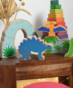 Lanka Kade Stegosaurus Dinosaur toy figure placed on a small wooden table with scenery pieces from the prehistoric play set in the background. These wooden toy figures are part of a huge range of Lanka Kade fair trade, wooden toys available here at Babipur.
