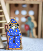 A Lanka Kade wooden toy wizard figure. The wizard has a blue coloured cloak with white and yellow stars and moon pattern withe a matching hat. The wizard is also holding a tall brown stick.