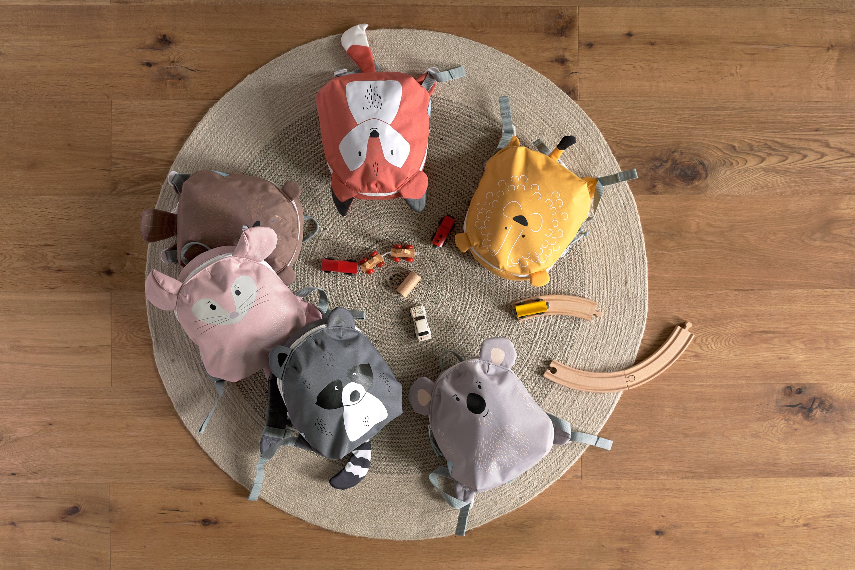 Six Lassig 'About Friends' animal backpacks for kids, laid out on a circular rug with some toys.