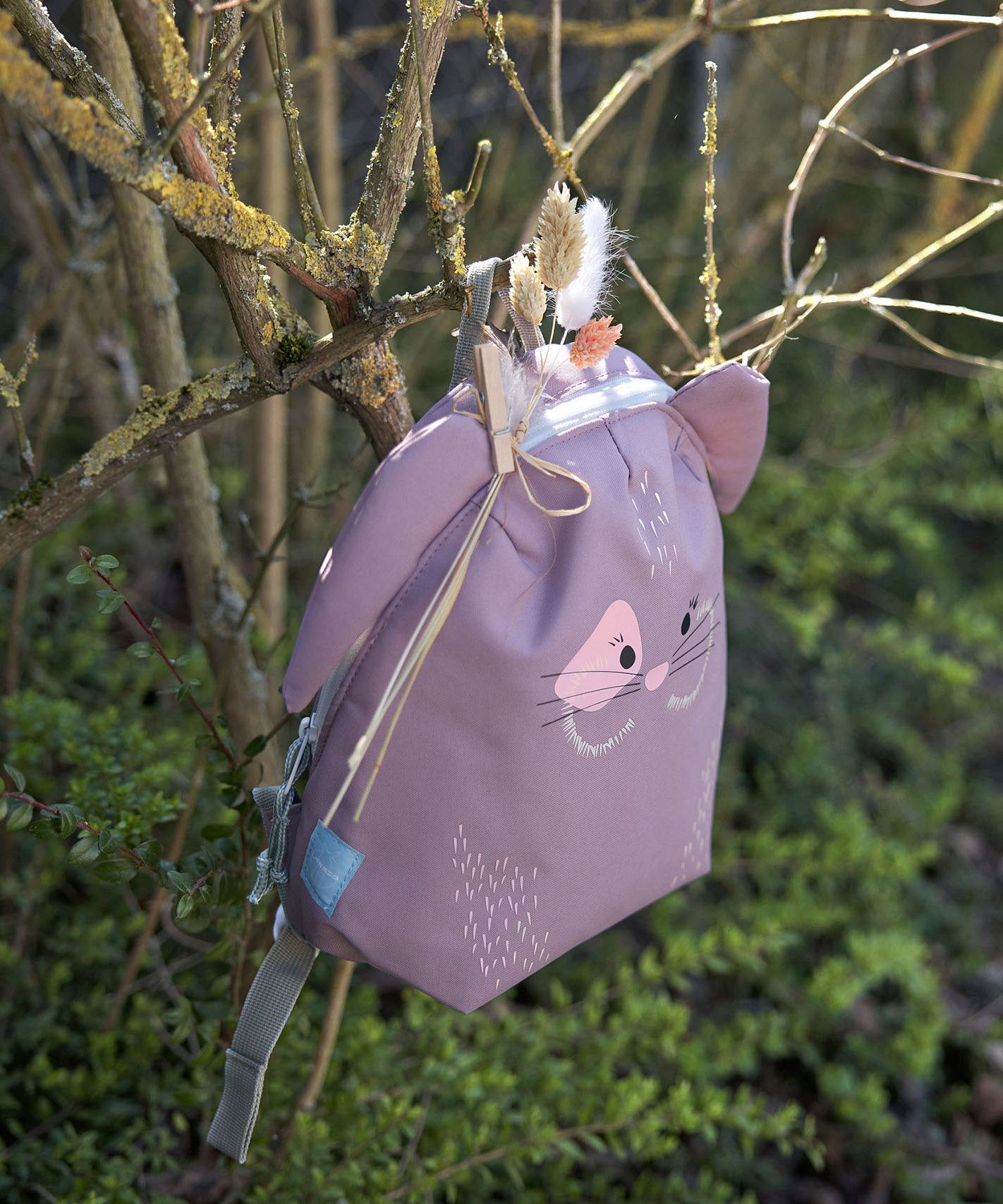 Lassig kids tiny bunny backpack hanging on a tree by the loop handle on the top with decorative reeds clipped on by a peg