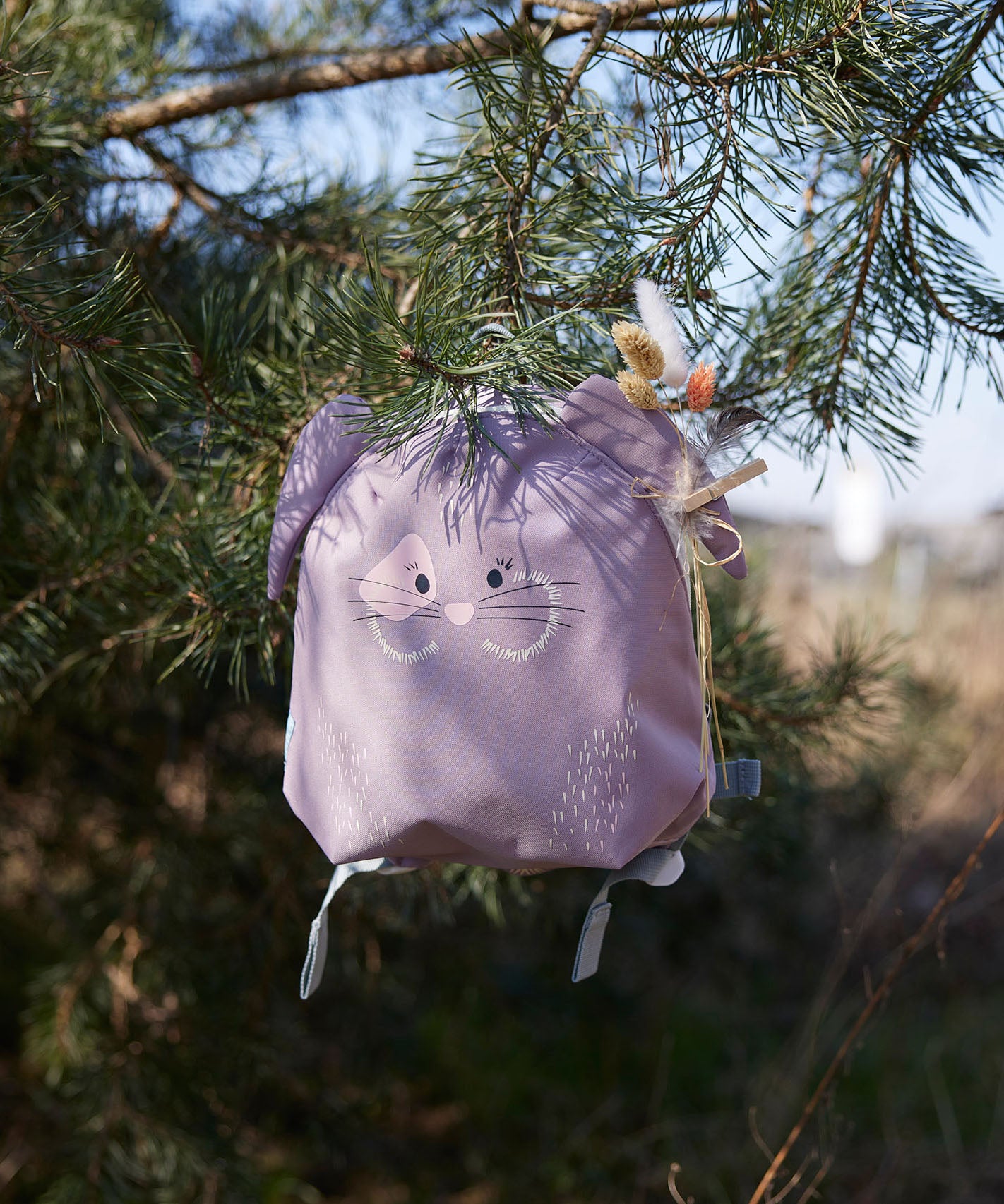 Lassig kids tiny bunny backpack hanging on a tree by the loop handle on the top with decorative reeds clipped on the ear by a peg
