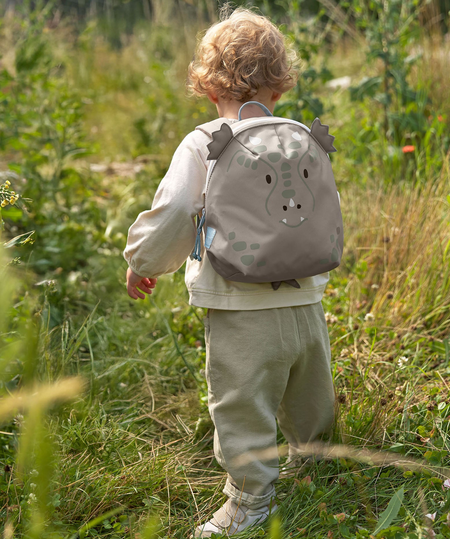 A child walking through a grassy gfield and wearing the Lassig kids tiny dragon backpack