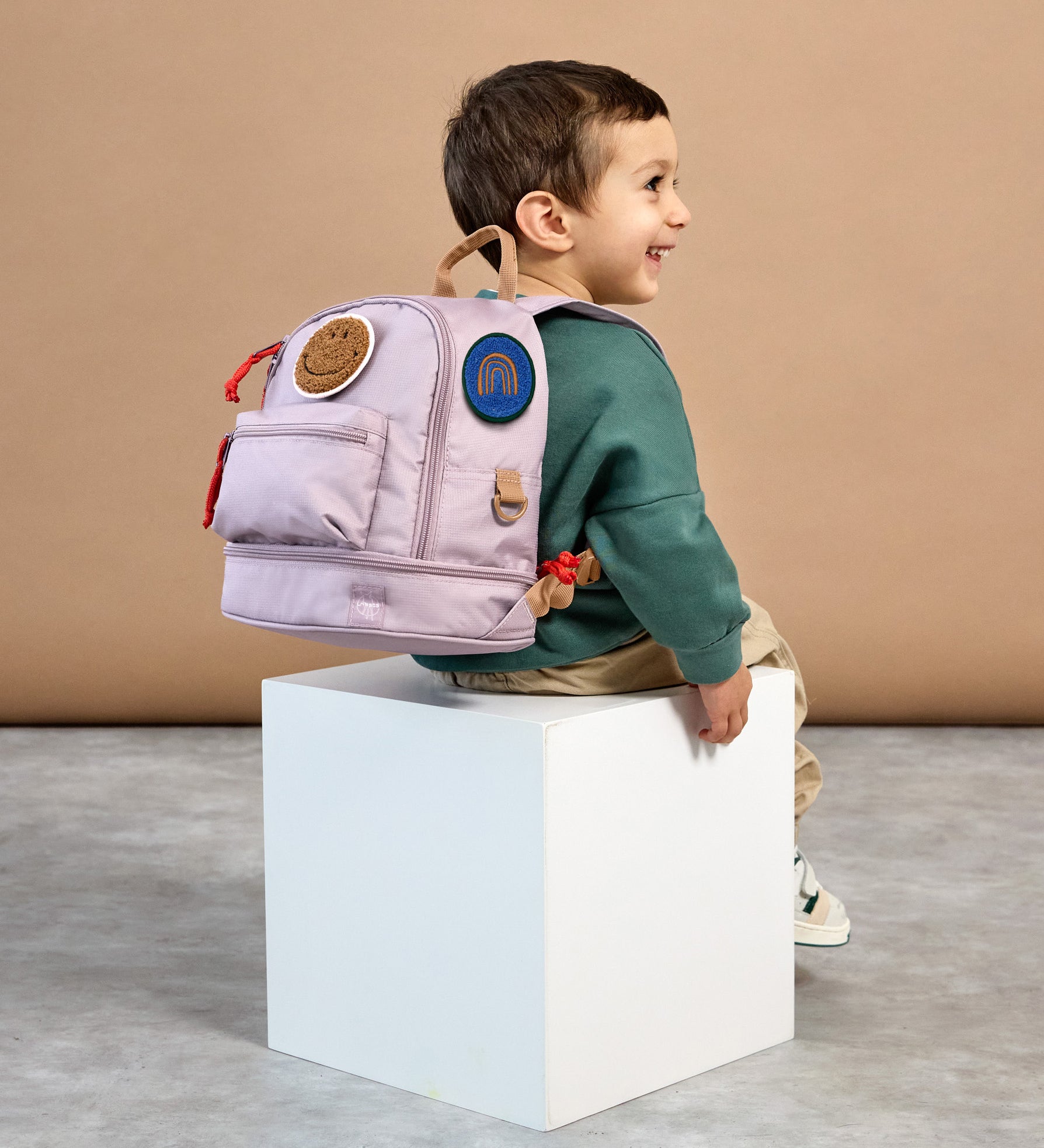 A little boy sat down, wearing the Lassig mini kids backpack in a lilac colour.