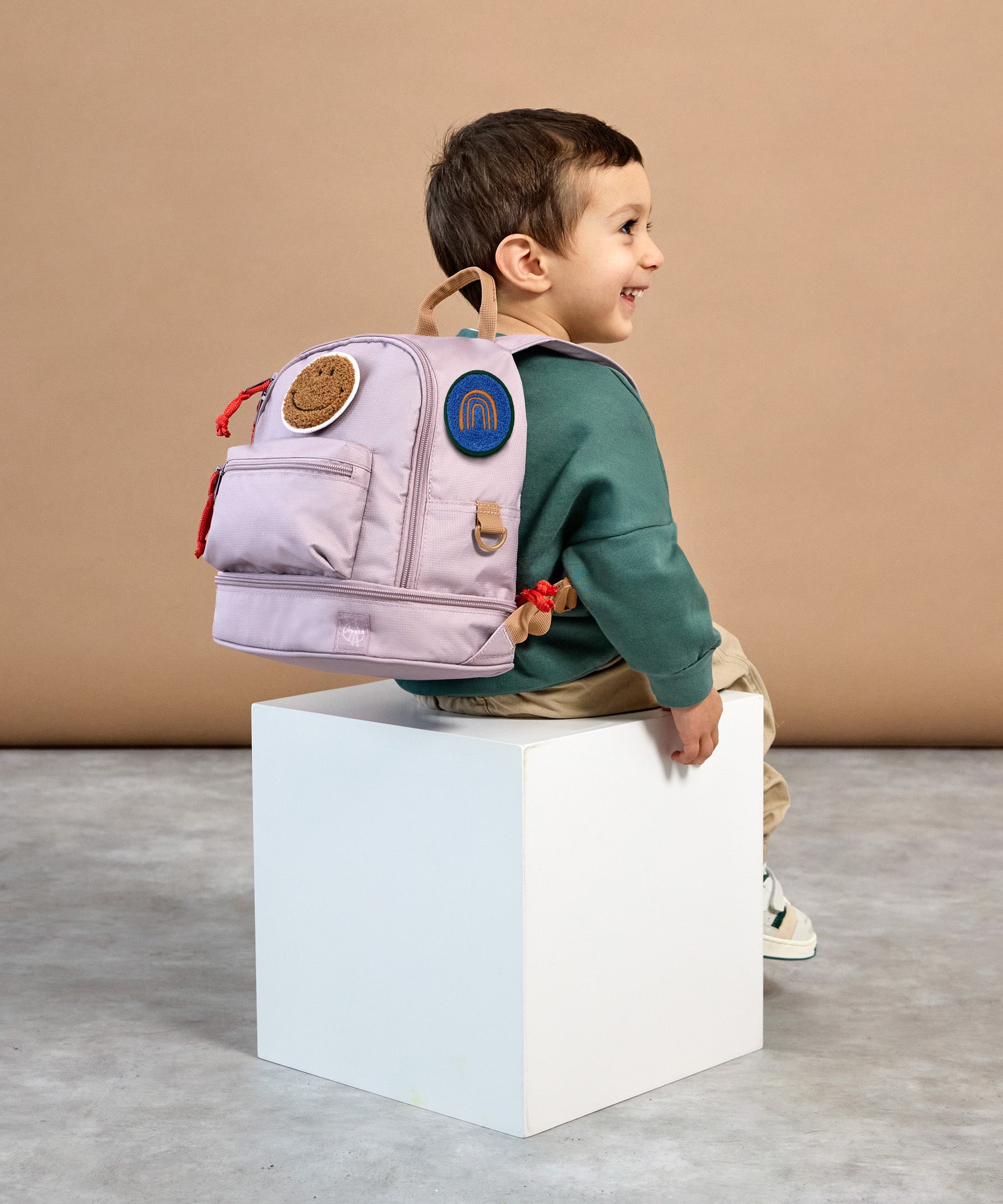 A little boy sat down, wearing the Lassig mini kids backpack in a lilac colour.