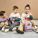 Three children sat together, one girl has a Lassig mini kids backpack in a lilac colour