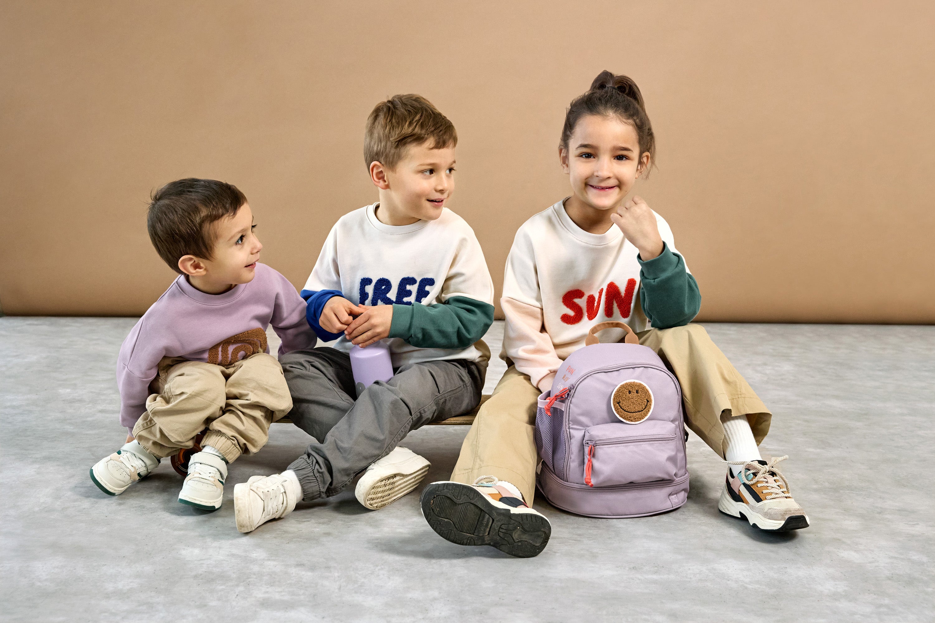 Three children sat together, one girl has a Lassig mini kids backpack in a lilac colour