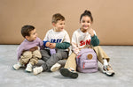 Three children sat together, one girl has a Lassig mini kids backpack in a lilac colour