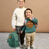 A boy holding the Lassig mini backpack for kids in a dark green colour, with his arm around a younger boy who's wearing the lilac version.